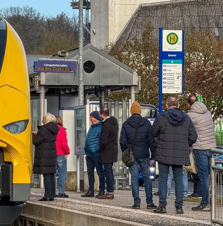 Fahrgäste warten an einer Haltestelle neben einem einfahrenden gelben Triebwagen